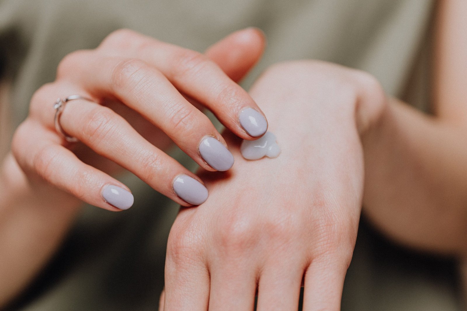 Close-up of a hand with gray-lilac nail polish applying a dollop of cream lotion to the back of the other hand